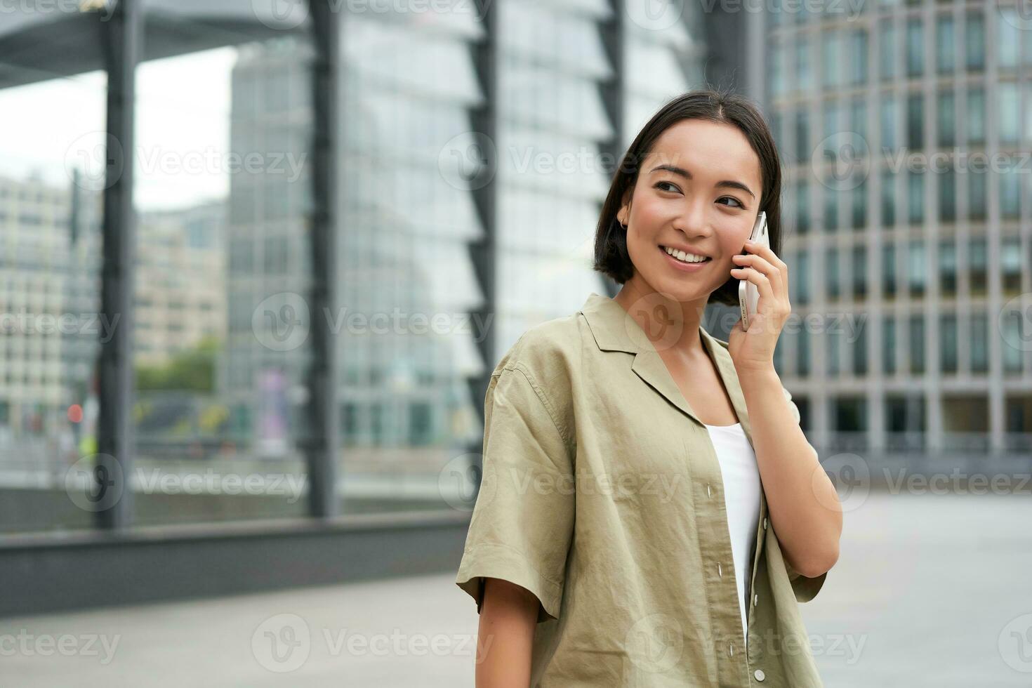 ritratto di asiatico ragazza sorridente, parlando su Telefono, fabbricazione un' chiamata, in piedi su strada vicino edificio e in attesa per qualcuno, risposta telefono foto