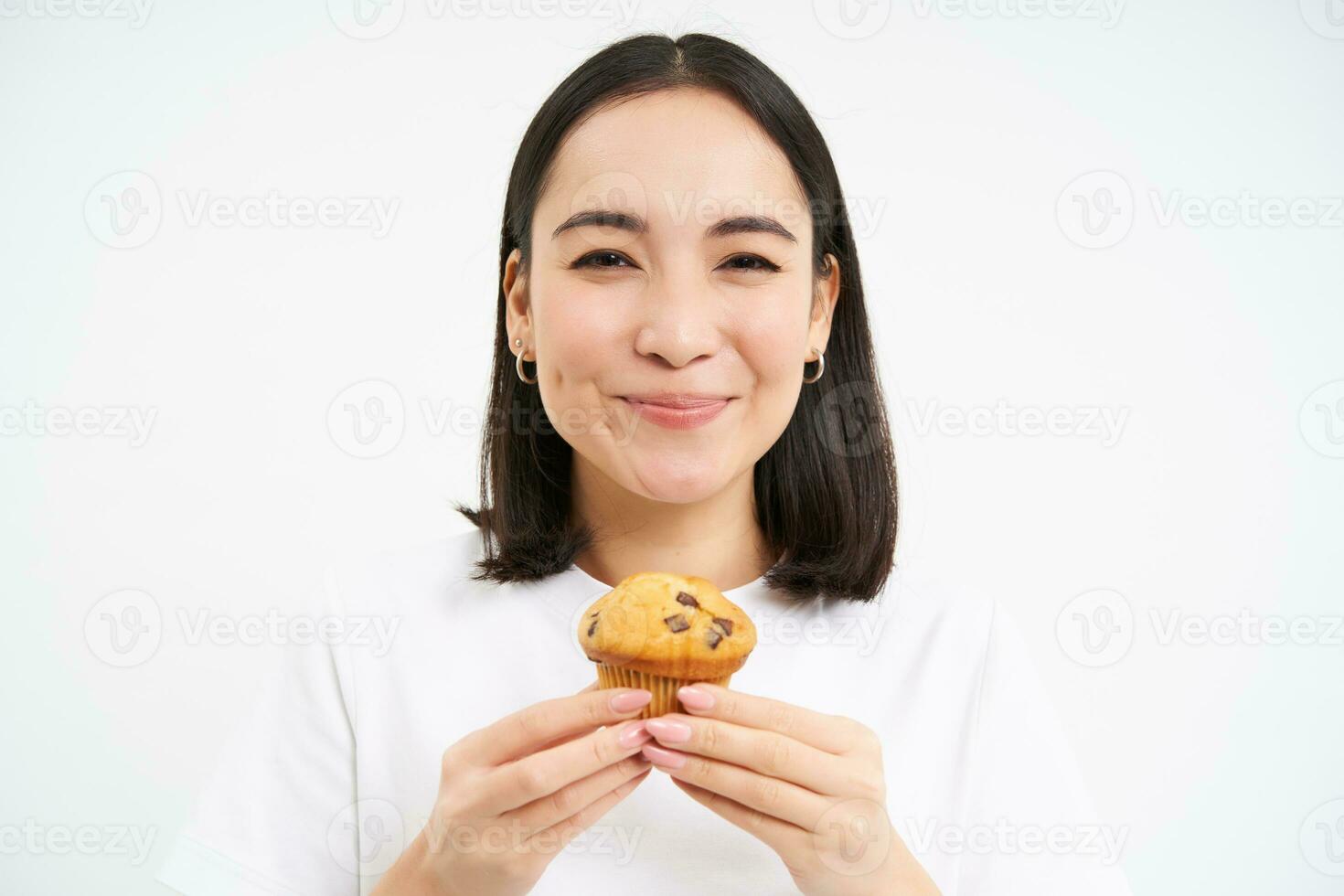 vicino su ritratto di asiatico donna piace Pasticcino, morsi gustoso Cupcake con contento indifferente Sorridi, bianca sfondo foto