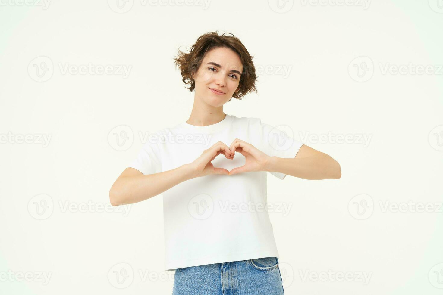 ritratto di brunetta ragazza Spettacoli cuore cartello, amore gesto, esprimere cura e caldo sentimenti, sta al di sopra di bianca sfondo foto