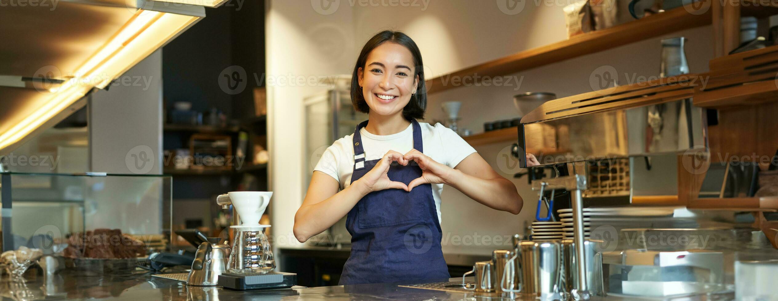sorridente asiatico ragazza barista, Spettacoli cuore cartello, gli amori fabbricazione caffè un servendo clienti, in piedi nel uniforme dietro a contatore, opera nel bar foto
