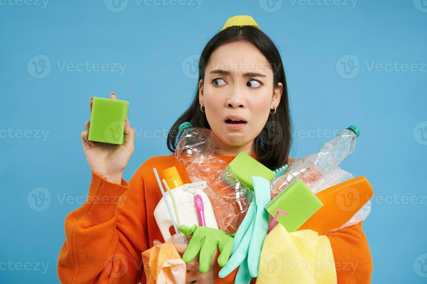ritratto di asiatico donna guardare disgustato a sporco spugna, Tenere riciclabile vuoto bottiglie, ordinamento spazzatura, blu sfondo foto