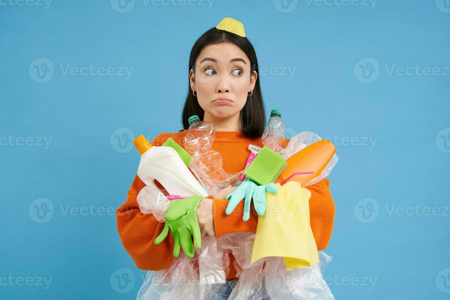 confuso giovane asiatico donna, Tenere plastica bottiglie, domestico spazzatura, ordinamento rifiuto per raccolta differenziata, blu sfondo foto