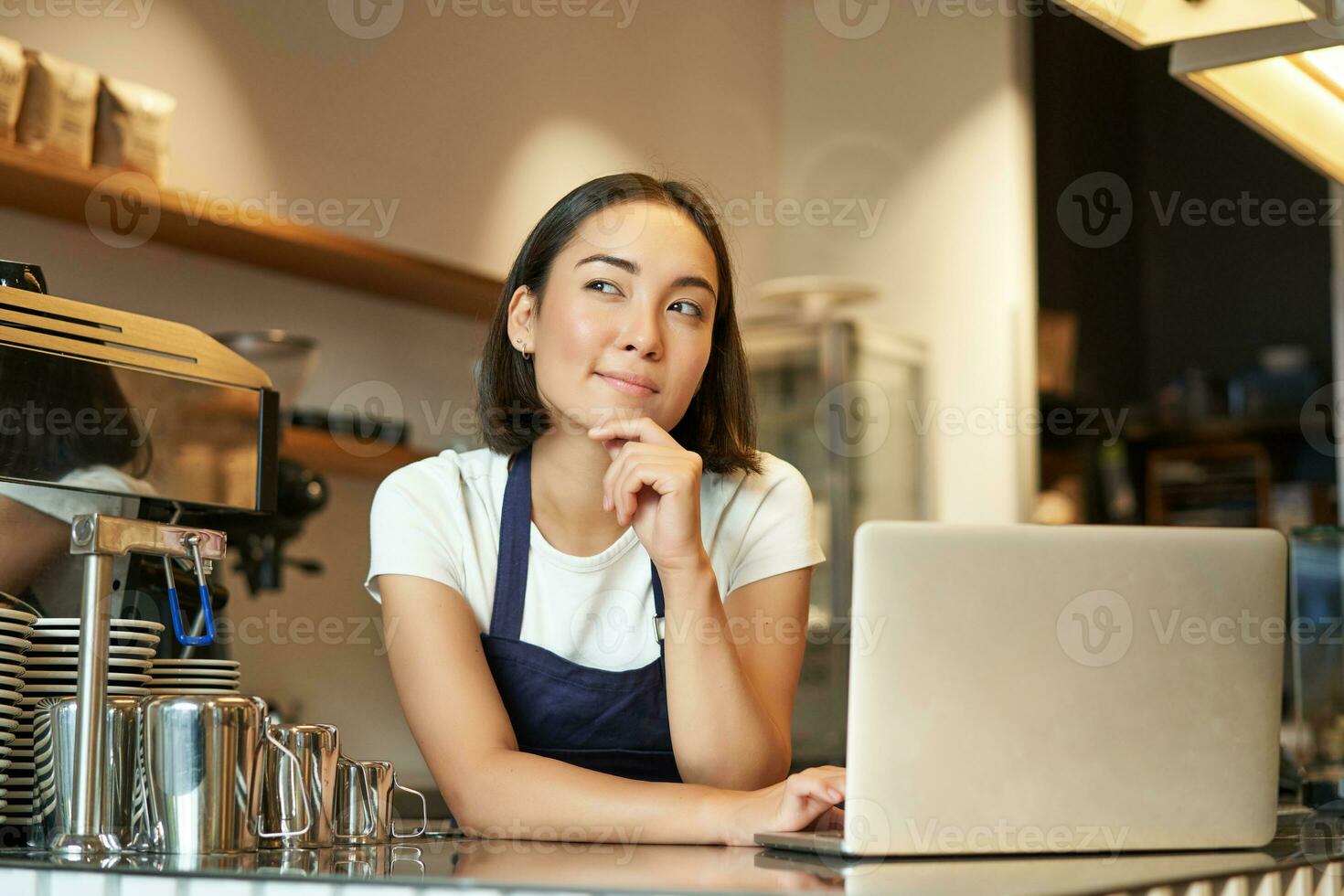 ritratto di ragazza barista con computer portatile, in piedi a contatore nel caffè negozio e pensiero, guardare curioso e pensiero foto