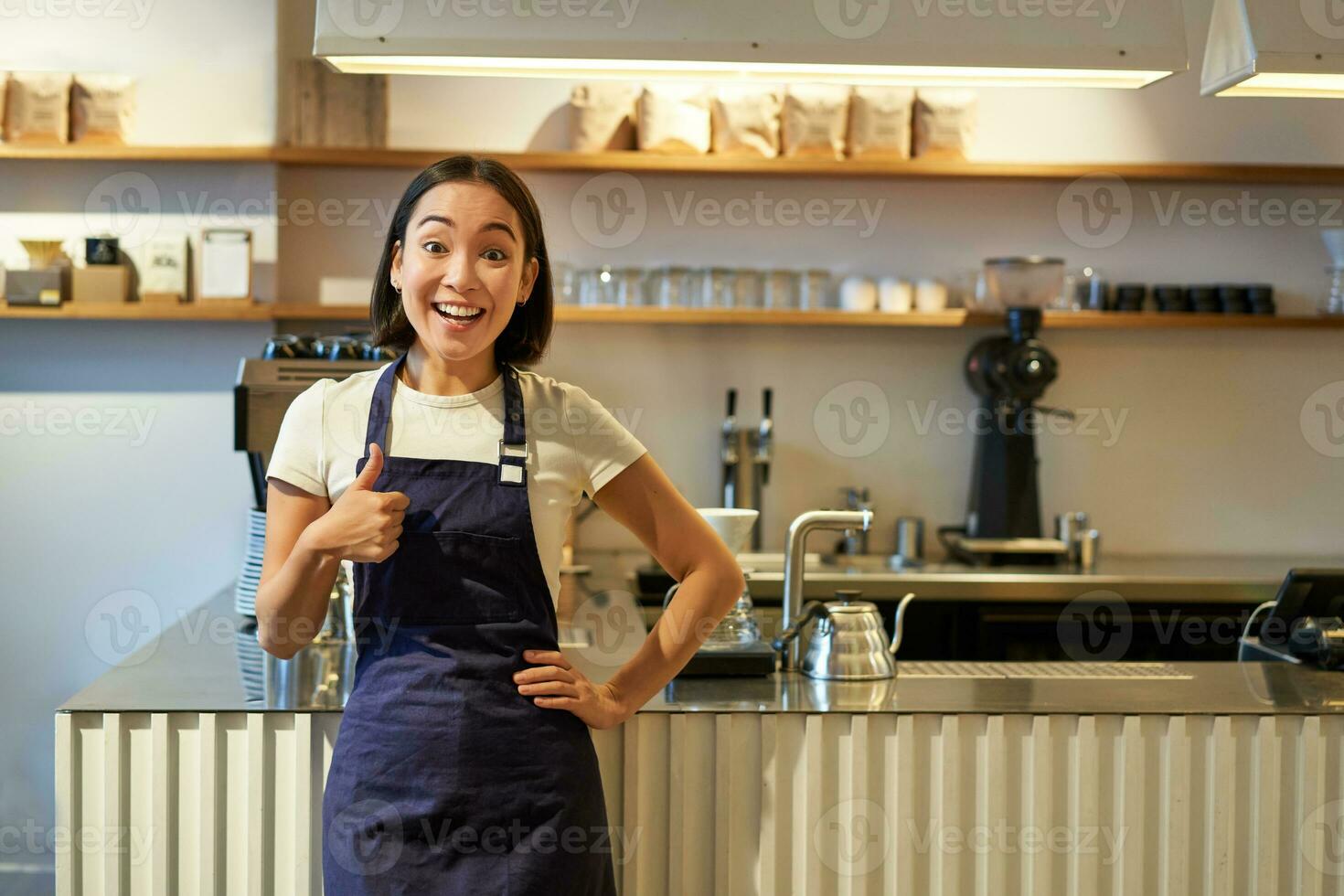 sorridente ragazza alunno Lavorando mezza giornata nel bar, barista Spettacoli pollici su, indossa grembiule, sta vicino caffè negozio contatore foto