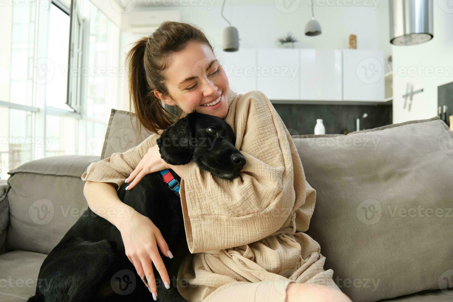 animali e stile di vita concetto. contento giovane donna nel accappatoio, abbracci sua cane su divano, coccolare cucciolo e sorridente foto