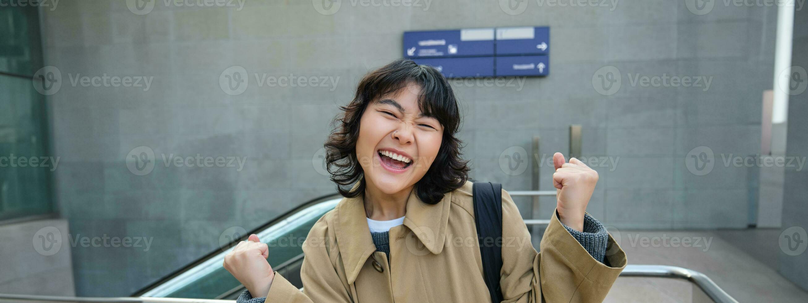 entusiasta asiatico ragazza sta su strada, fa evviva celebrazione gesto, dice sì, trionfante, vincente e godendo Questo sensazione, in piedi solo all'aperto foto