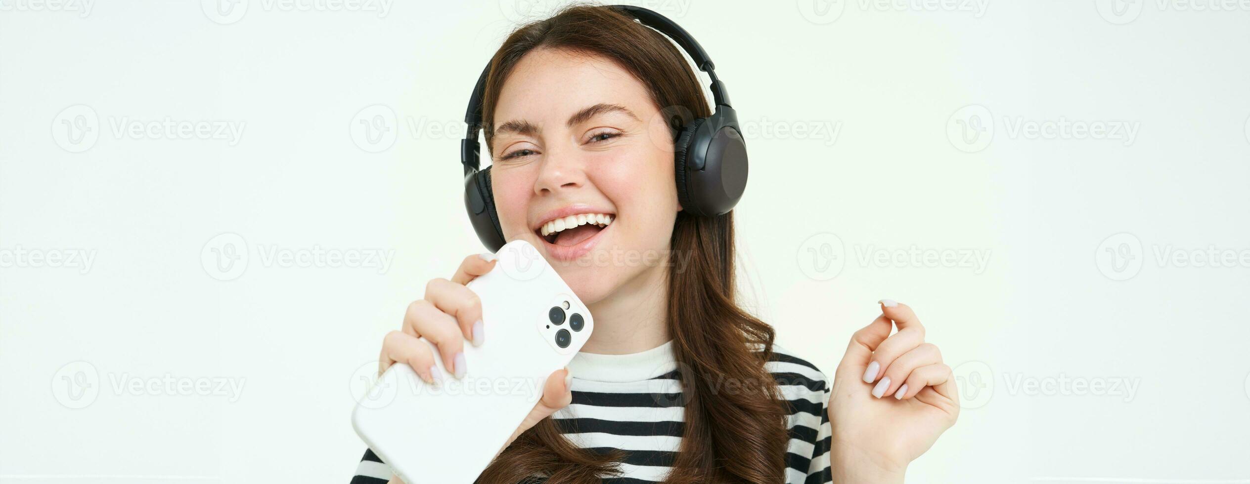 tecnologia e sociale media. ritratto di contento giovane donna cantando karaoke, giocando musica App su smartphone, indossare senza fili cuffia, in piedi al di sopra di bianca sfondo foto