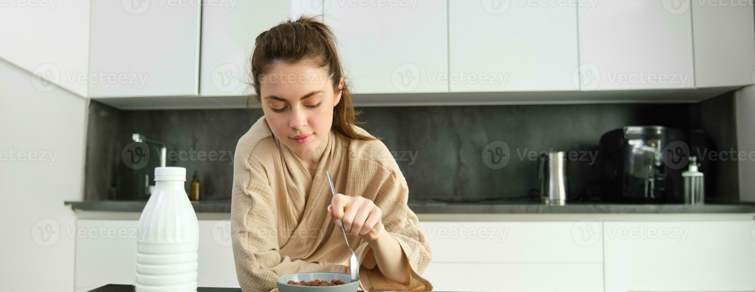 Immagine di contento giovane donna mangiare cereali a casa con latte, avendo sua colazione, indossare accappatoio, seduta nel cucina solo foto