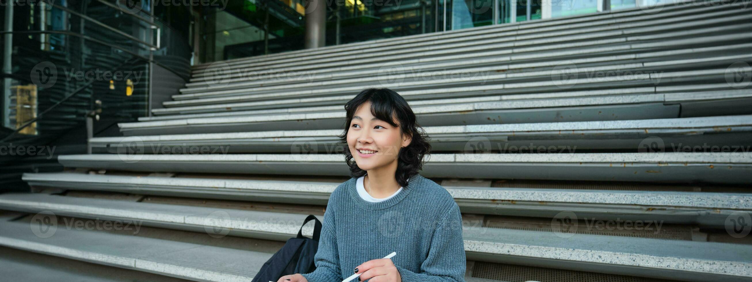 sorridente ragazza, grafico progettista, utilizzando digitale tavoletta e penna attrezzo per disegno, fa casa Incarico per Università, si siede su le scale foto