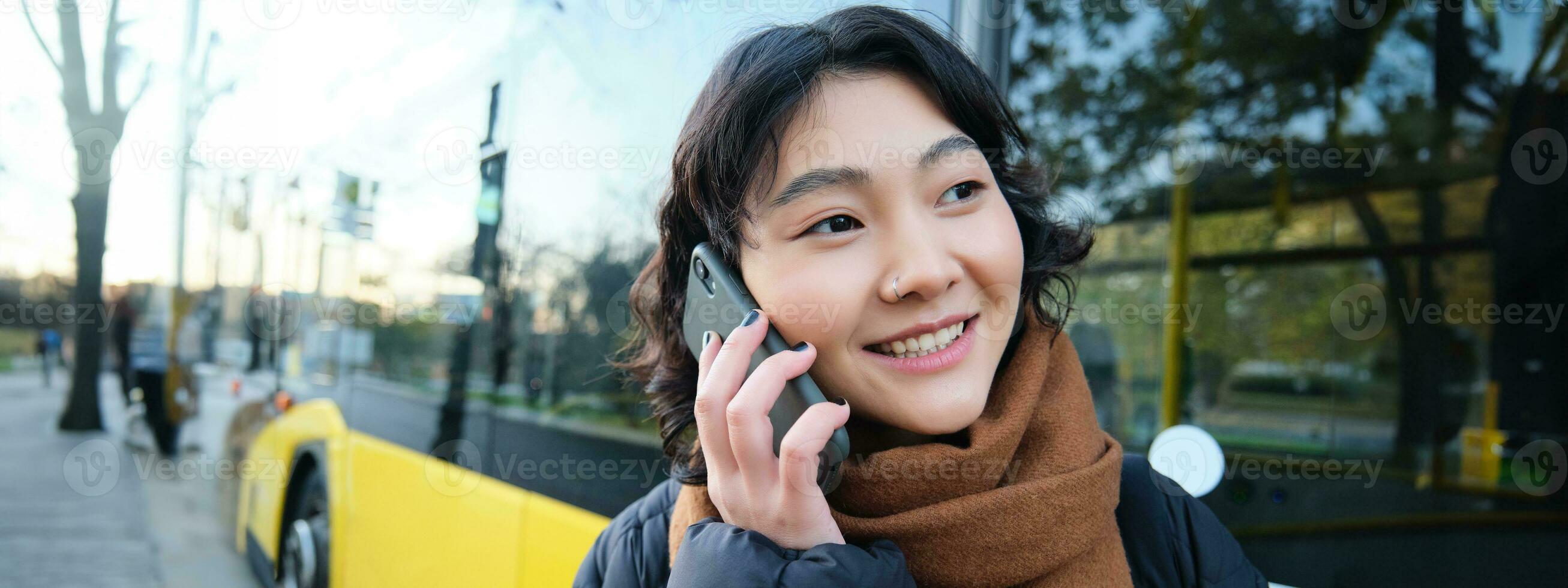 cellulare tecnologia e persone concetto. elegante asiatico ragazza parla su mobile Telefono, fa un' telefono chiamata, sta vicino autobus fermare e ha conversazione foto