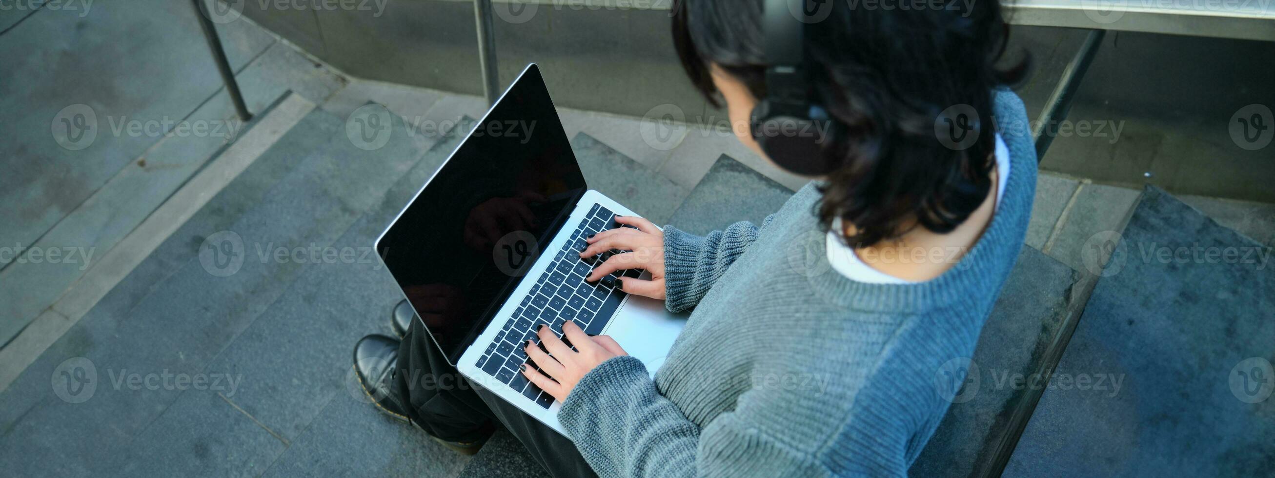 vicino su ritratto di ragazza, alunno lavori su il computer portatile e ascolta musica nel cuffia. vuoto computer schermo e mani digitando su tastiera foto