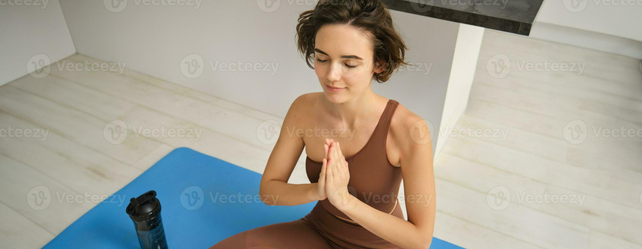giovane salutare bellissimo donna nel gli sport superiore e ghette praticante yoga a casa, seduta nel loto posizione su yoga stuoia, meditando, sorridente, rilassato, occhi Chiuso, meditazione concetto foto