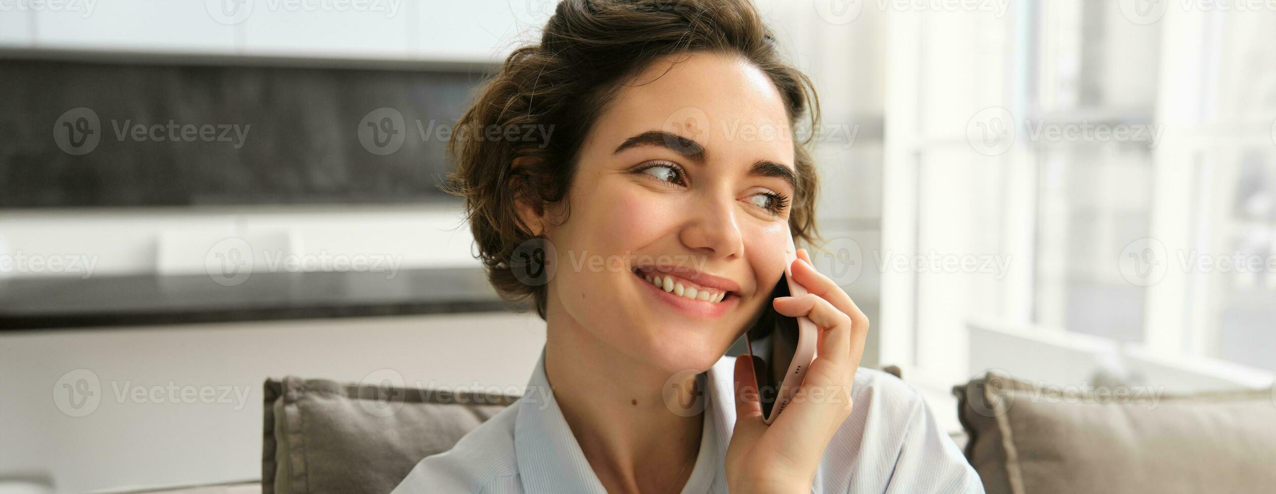 vicino su ritratto di sorridente bellissimo donna, parlando su mobile Telefono, chat su smartphone, chiamate qualcuno, si siede a casa foto