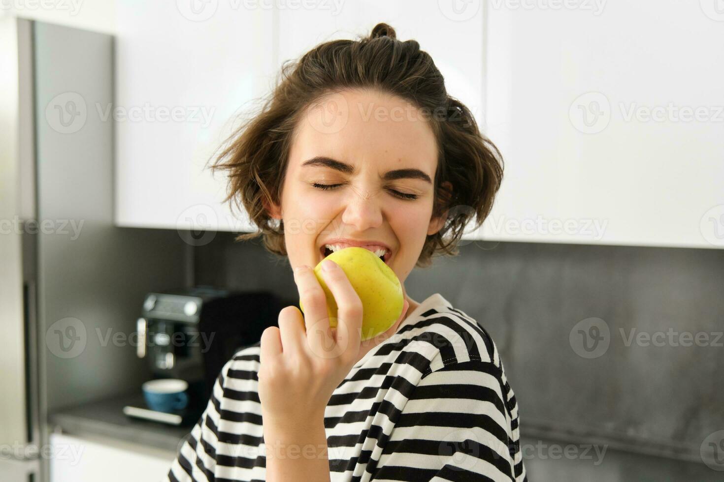 vicino su ritratto di giovane brunetta donna pungente un Mela con piacere, ha lieta Sorridi su sua viso, in piedi nel il cucina, avendo salutare merenda per il pranzo, mangiare frutta foto