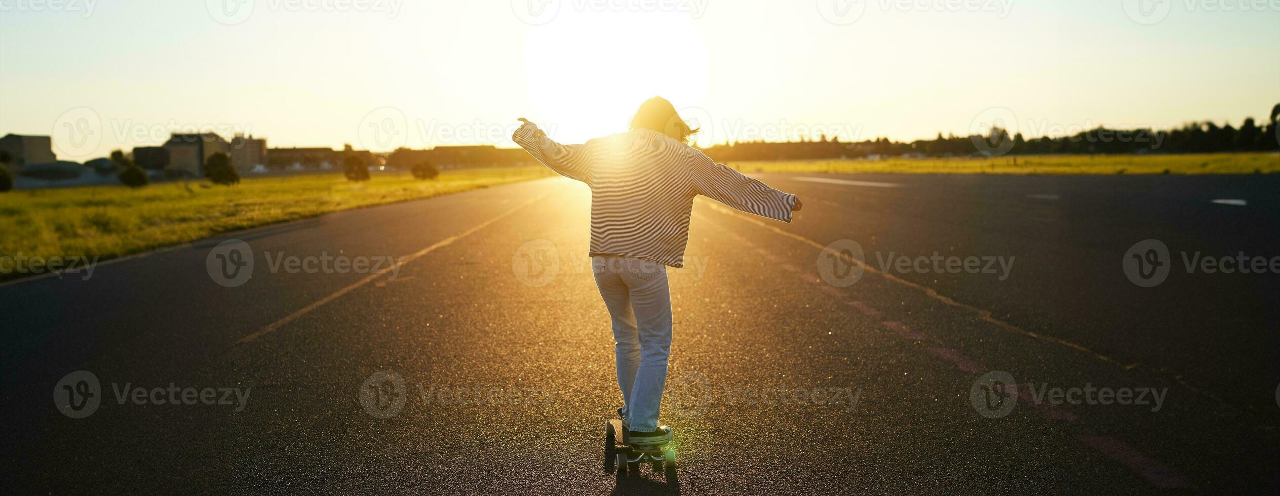 adolescente ragazza sensazione contento su longboard. contento giovane pattinatore equitazione sua skateboard con mani diffusione di lato, sensazione libertà, andando in direzione il sole foto