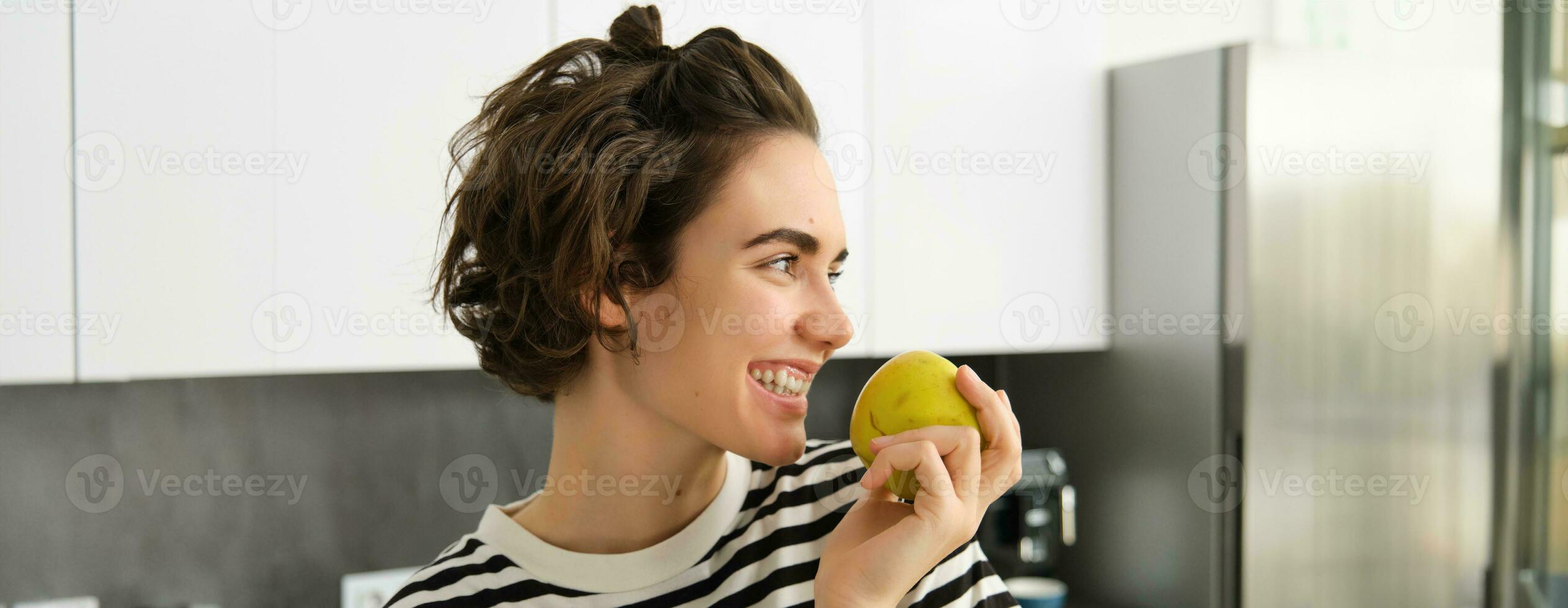 vicino su ritratto di carino giovane moderno donna, mangiare verde Mela e guardare a parte, sorridente mentre pungente un' frutta, avendo un' merenda nel il cucina foto