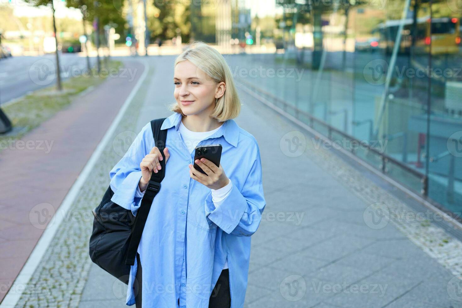 ritratto di biondo giovane donna, utilizzando navigazione carta geografica App su smartphone, a piedi lungo strada, utilizzando indicazioni su Telefono, Tenere zaino foto