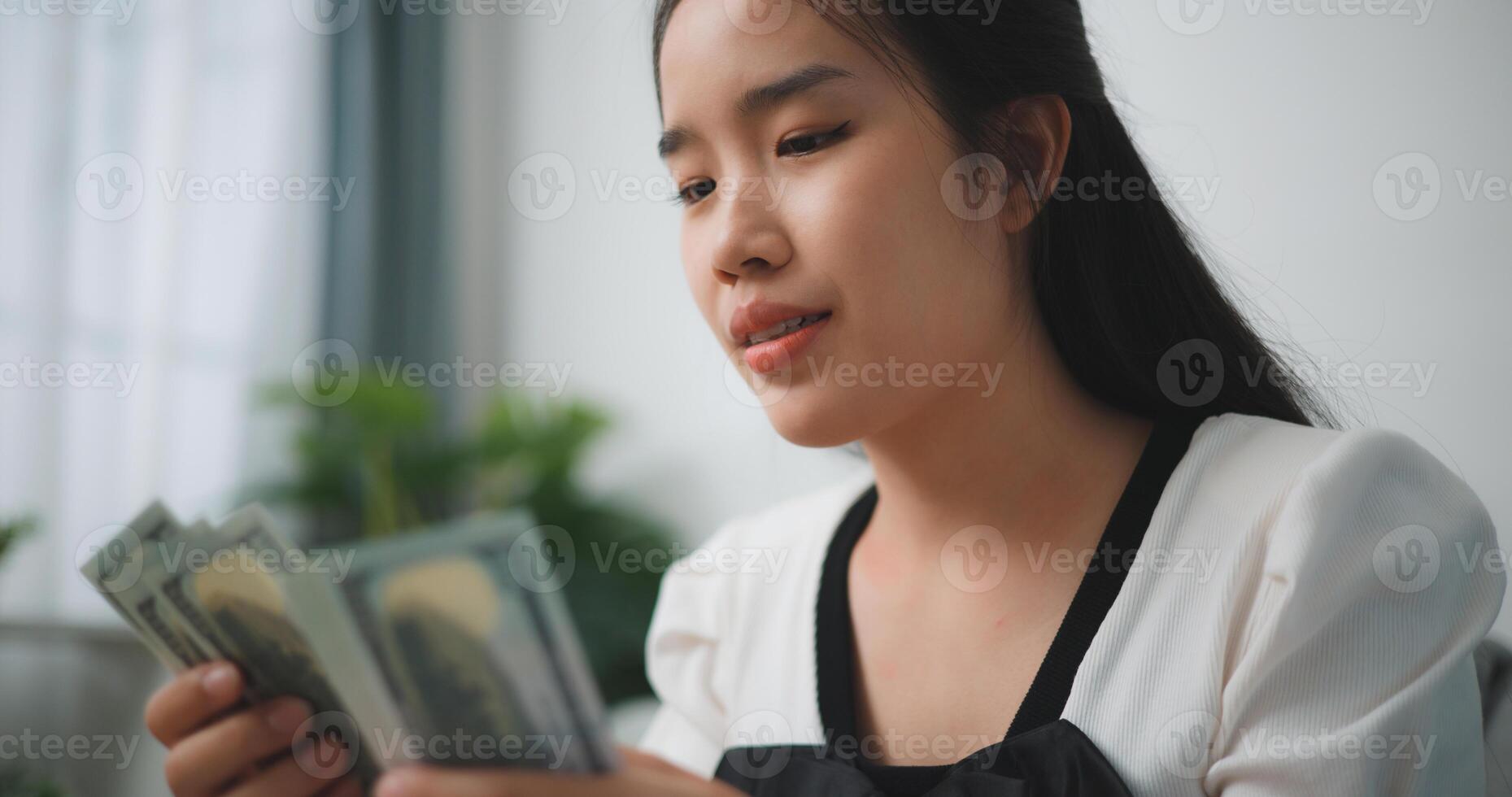 ritratto di contento giovane asiatico donna conta denaro contante dollaro fatture mentre seduta nel vivente camera a a casa, contando pila denaro contante i soldi dollaro foto