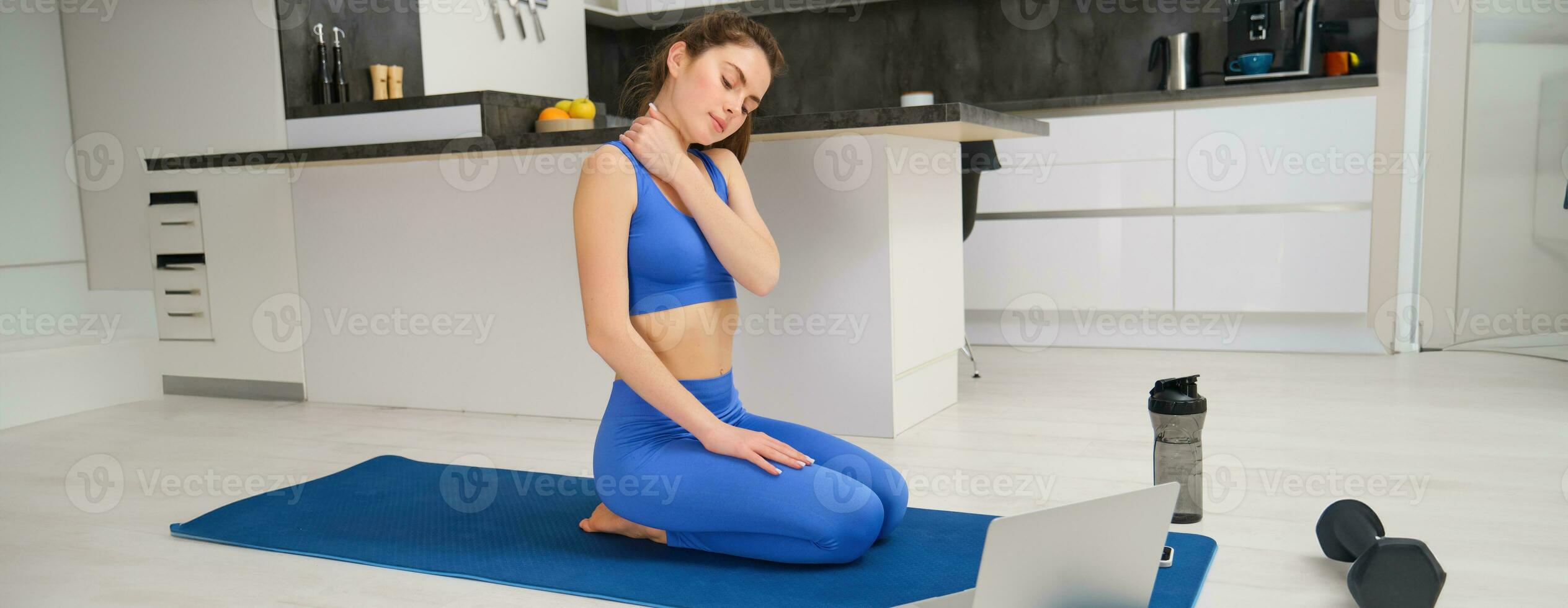 Immagine di calma e rilassato donna, seduta su yoga stuoia con computer portatile, massaggio sua collo, riscaldamento corpo prima allenarsi addestramento, casa fitness sessione foto