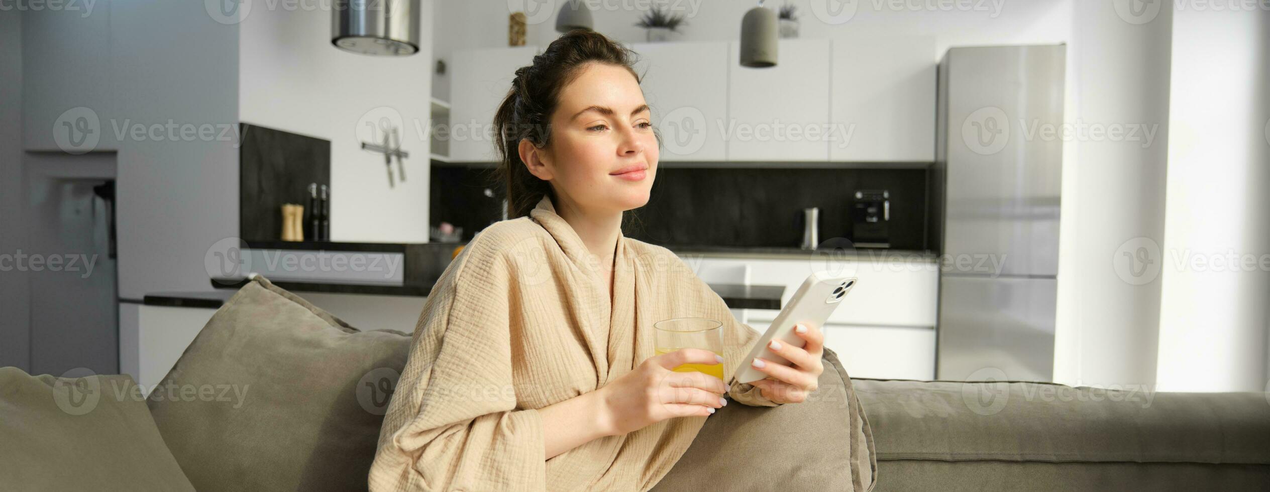 bellissimo giovane donna riposo su divano, godendo sua mattina a casa, Tenere bicchiere di arancia succo e smartphone, guardare al di fuori finestra foto