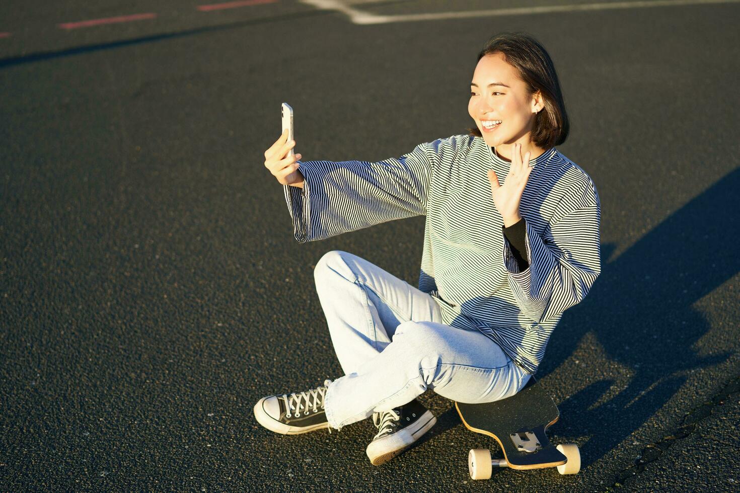 positivo adolescente asiatico ragazza prende autoscatto, video chat su mobile Telefono app, si siede su skateboard e record video blog foto