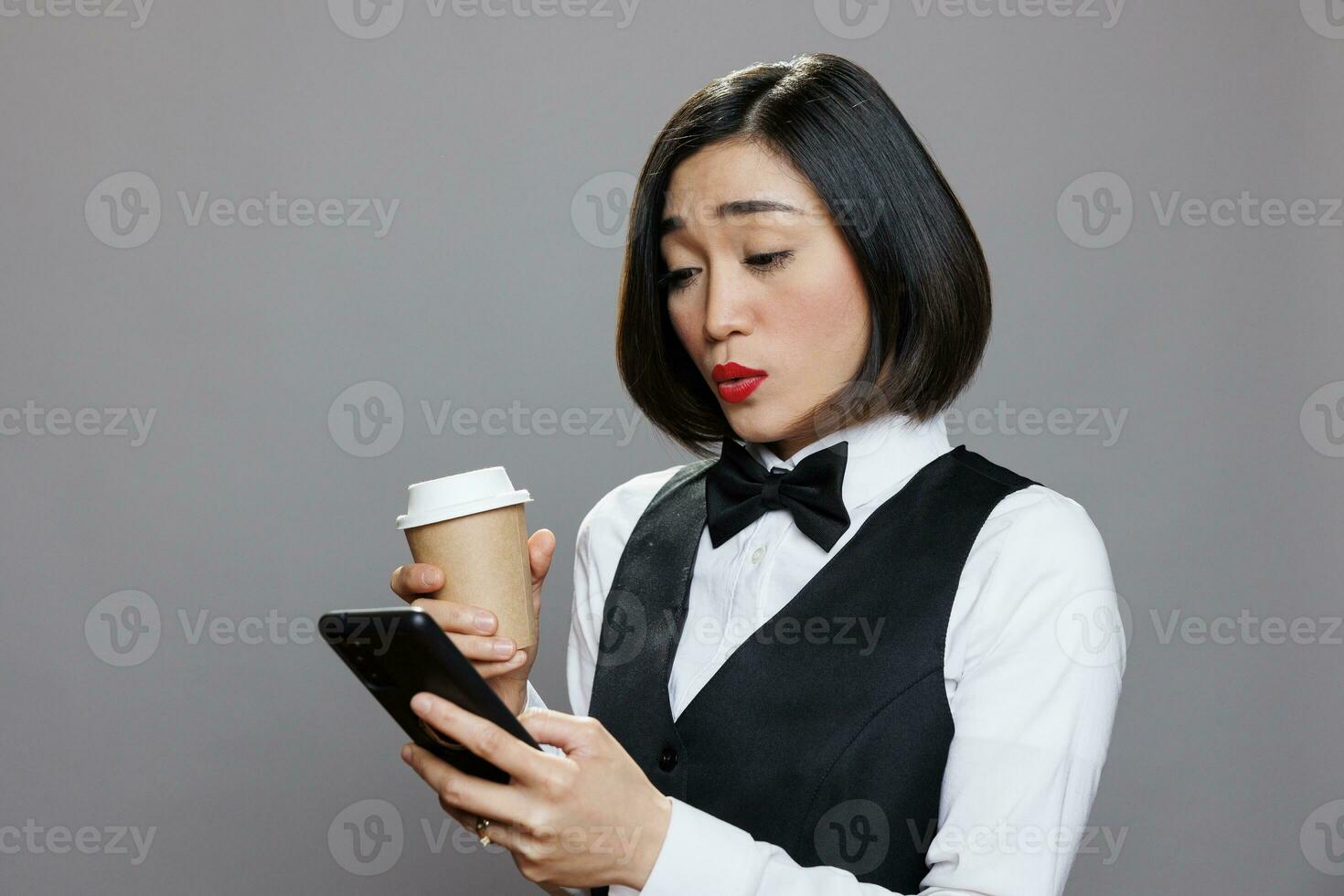 asiatico donna nel cameriera uniforme invio Messaggio su mobile Telefono durante caffè rompere. giovane addetto alla reception parlando su mobile Telefono sociale media e potabile caldo bevanda per partire foto