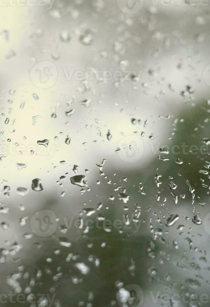 un' foto di pioggia gocce su il finestra bicchiere con un' sfocato Visualizza di il fioritura verde alberi. astratto Immagine mostrando nuvoloso e piovoso tempo metereologico condizioni