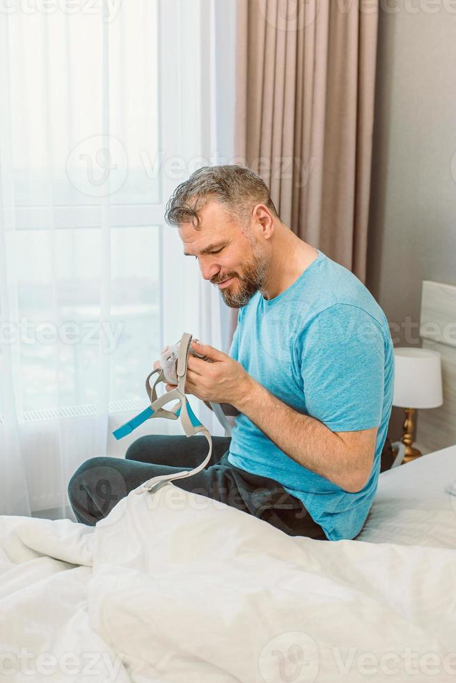 uomo felice riposato con problemi respiratori cronici dopo aver usato la macchina cpap seduto sul letto in camera da letto. assistenza sanitaria, cpap, terapia dell'apnea ostruttiva del sonno, concetto di russamento foto