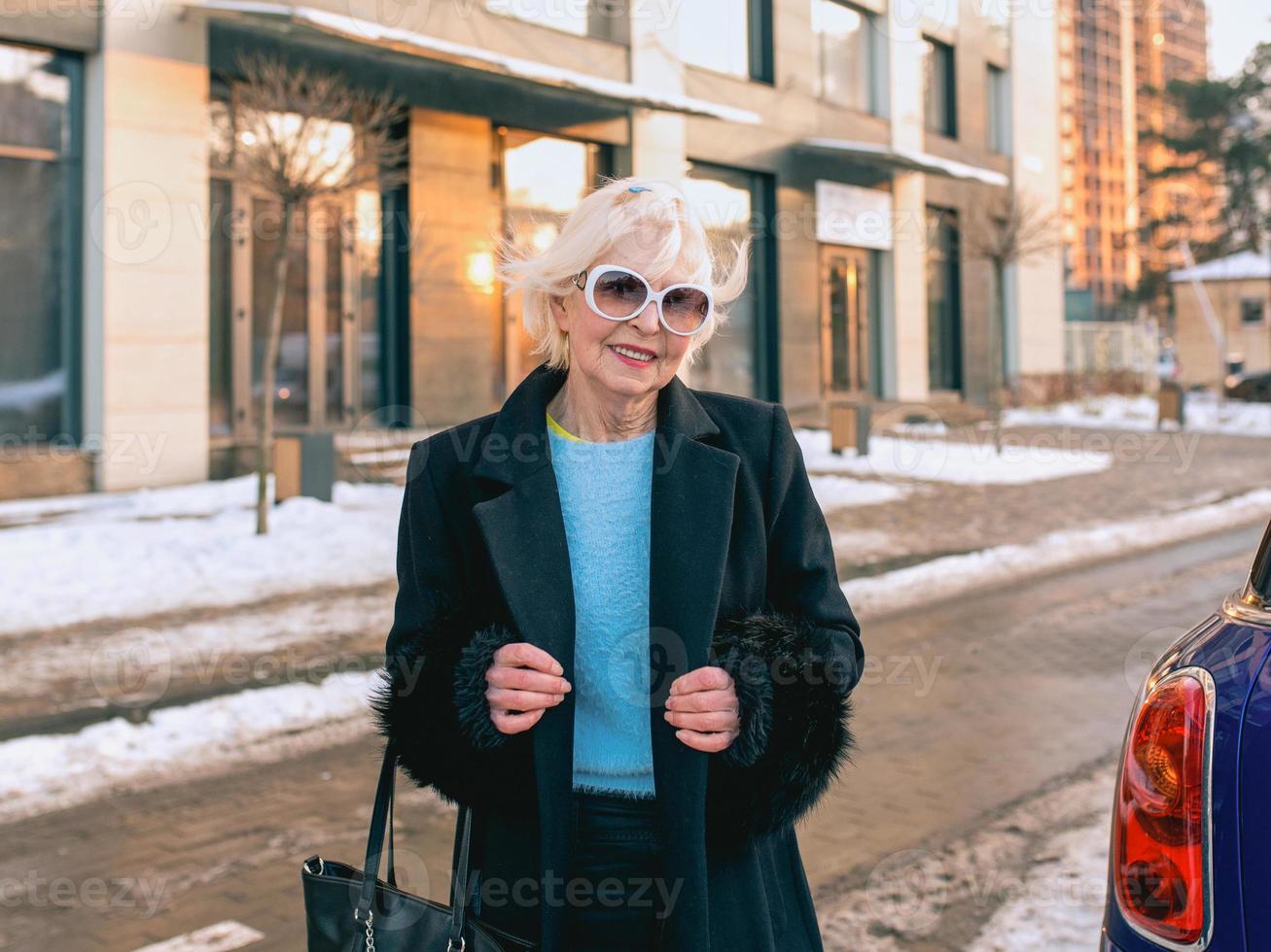 donna alla moda anziana in elegante cappotto nero e borsa a mano che cammina dalla sua auto all'appuntamento di lavoro. business, stile, concetto anti età foto