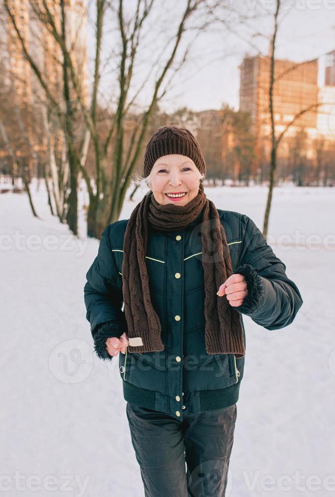 donna anziana in cappello e giacca sportiva che fa jogging nel parco invernale di neve. inverno, età, sport, attività, concetto di stagione foto