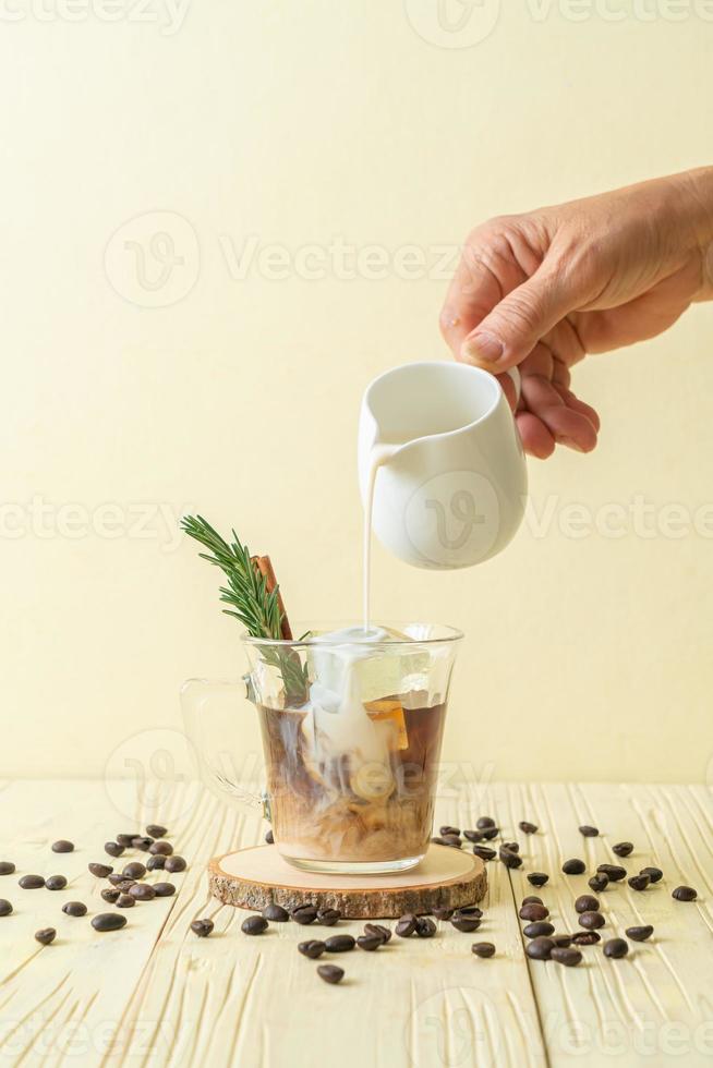 versare il latte nel bicchiere da caffè nero con cubetto di ghiaccio, cannella e rosmarino foto