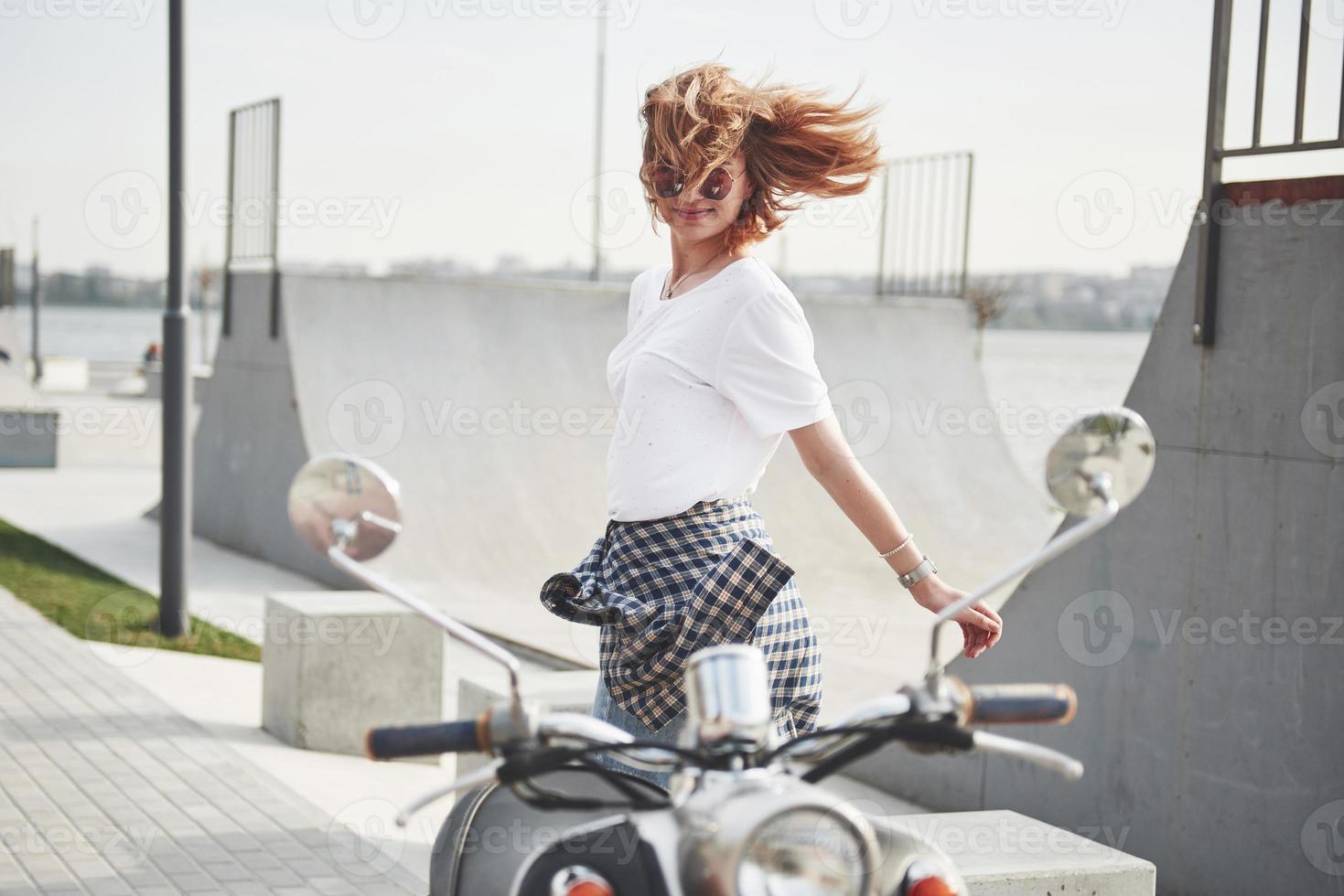 ritratto di una bella ragazza hipster seduta su uno scooter retrò nero, sorridente in posa e godersi il caldo sole primaverile. foto