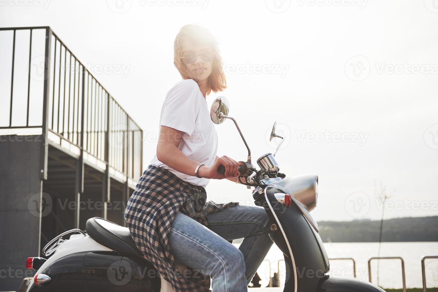 ritratto di una bella ragazza hipster seduta su uno scooter retrò nero, sorridente in posa e godersi il caldo sole primaverile. foto