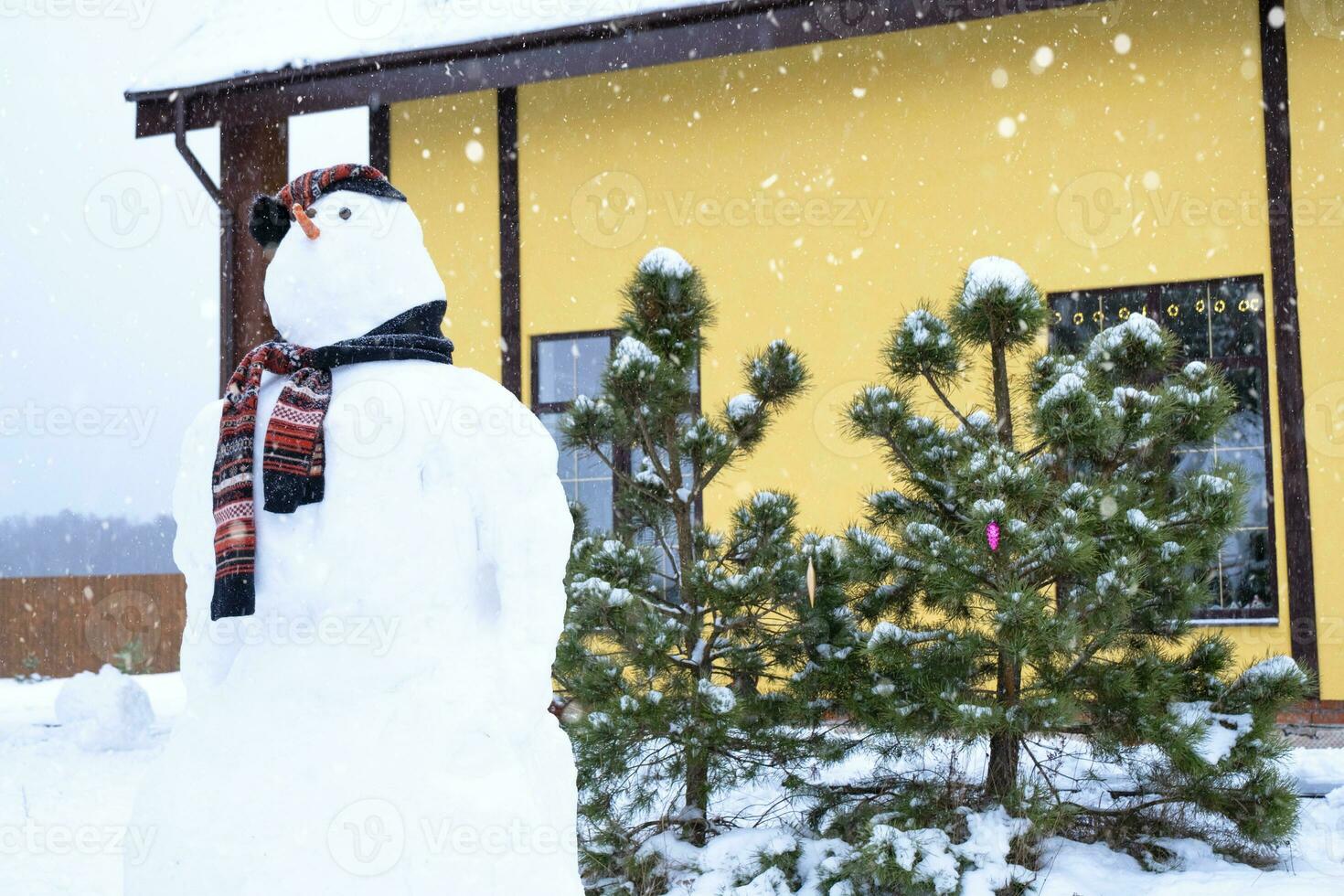 divertente pupazzo di neve nel un' cappello e sciarpa su il sfondo di un' giallo Casa nel il cortile. inverno, inverno intrattenimento, nevicata foto