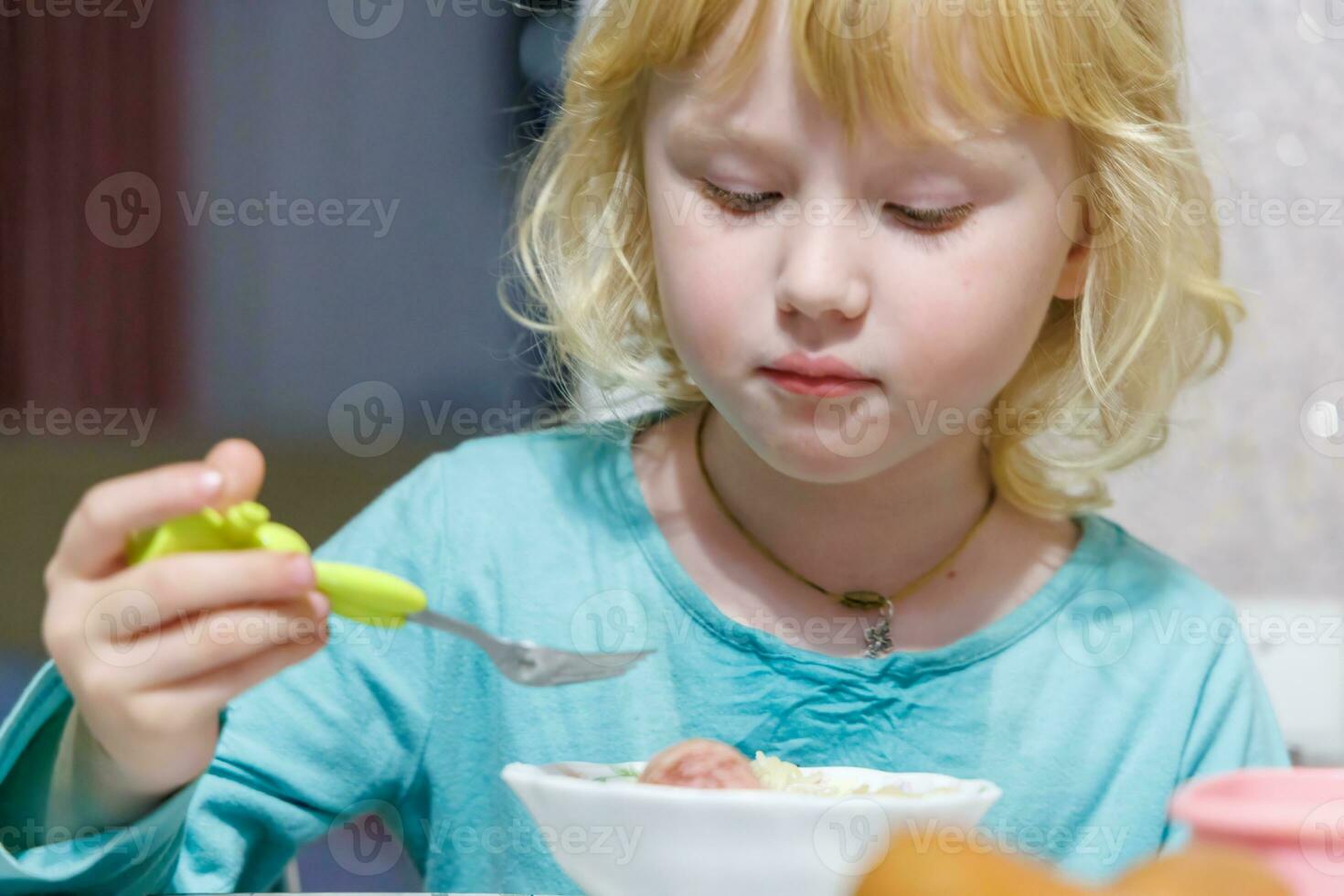 un' poco ragazza ha prima colazione a casa spaghetti con salsicce. poco bionda ragazza mangiare cena con forchetta a tavolo foto