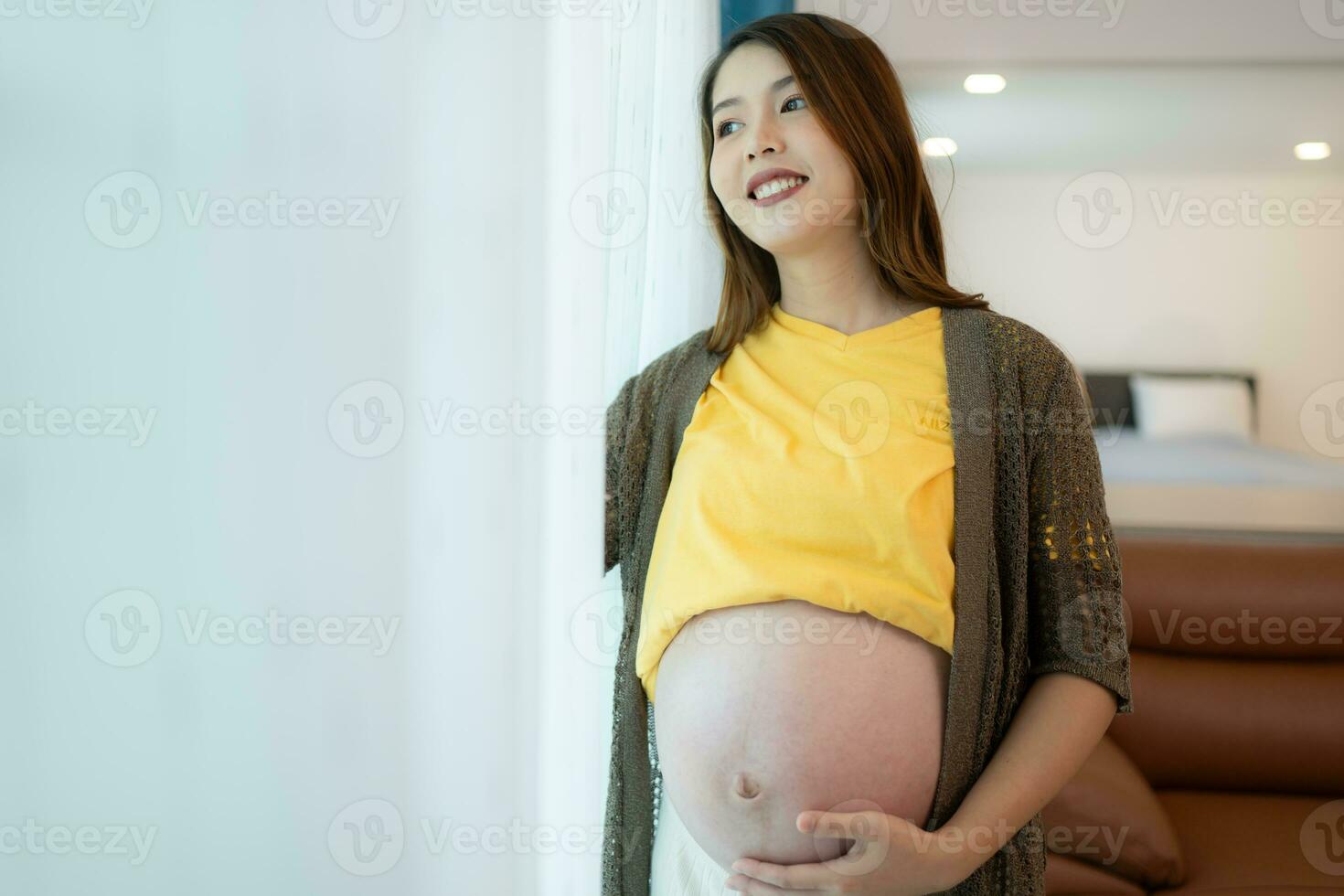 bellissimo incinta donna in piedi vicino finestra a casa e Tenere sua pancia foto