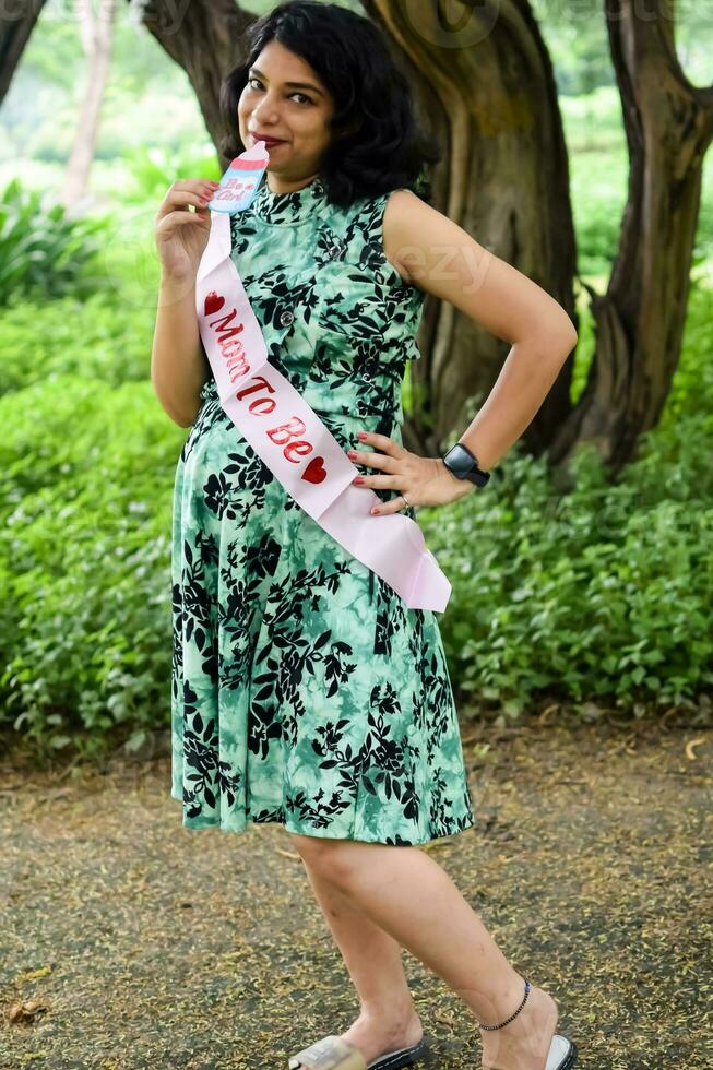 un' incinta indiano signora pose per all'aperto gravidanza sparare e mani su gonfiarsi, indiano incinta donna mette sua mano su sua stomaco con un' maternità vestito a società parco, incinta al di fuori maternità sparare foto