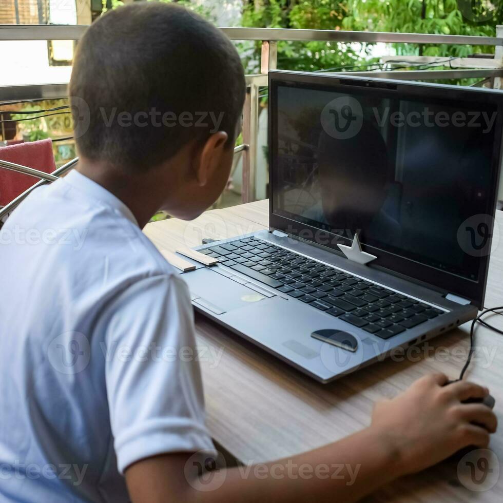 poco ragazzo seduta a tavolo utilizzando il computer portatile per in linea classe nel grado 1, bambino studiando su il computer portatile a partire dal casa per distanza apprendimento in linea formazione scolastica, scuola ragazzo bambini stile di vita concetto foto