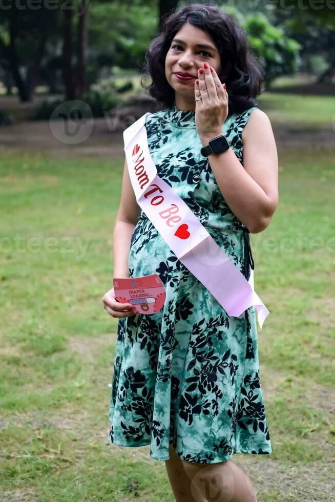 un' incinta indiano signora pose per all'aperto gravidanza sparare e mani su gonfiarsi, indiano incinta donna mette sua mano su sua stomaco con un' maternità vestito a società parco, incinta al di fuori maternità sparare foto