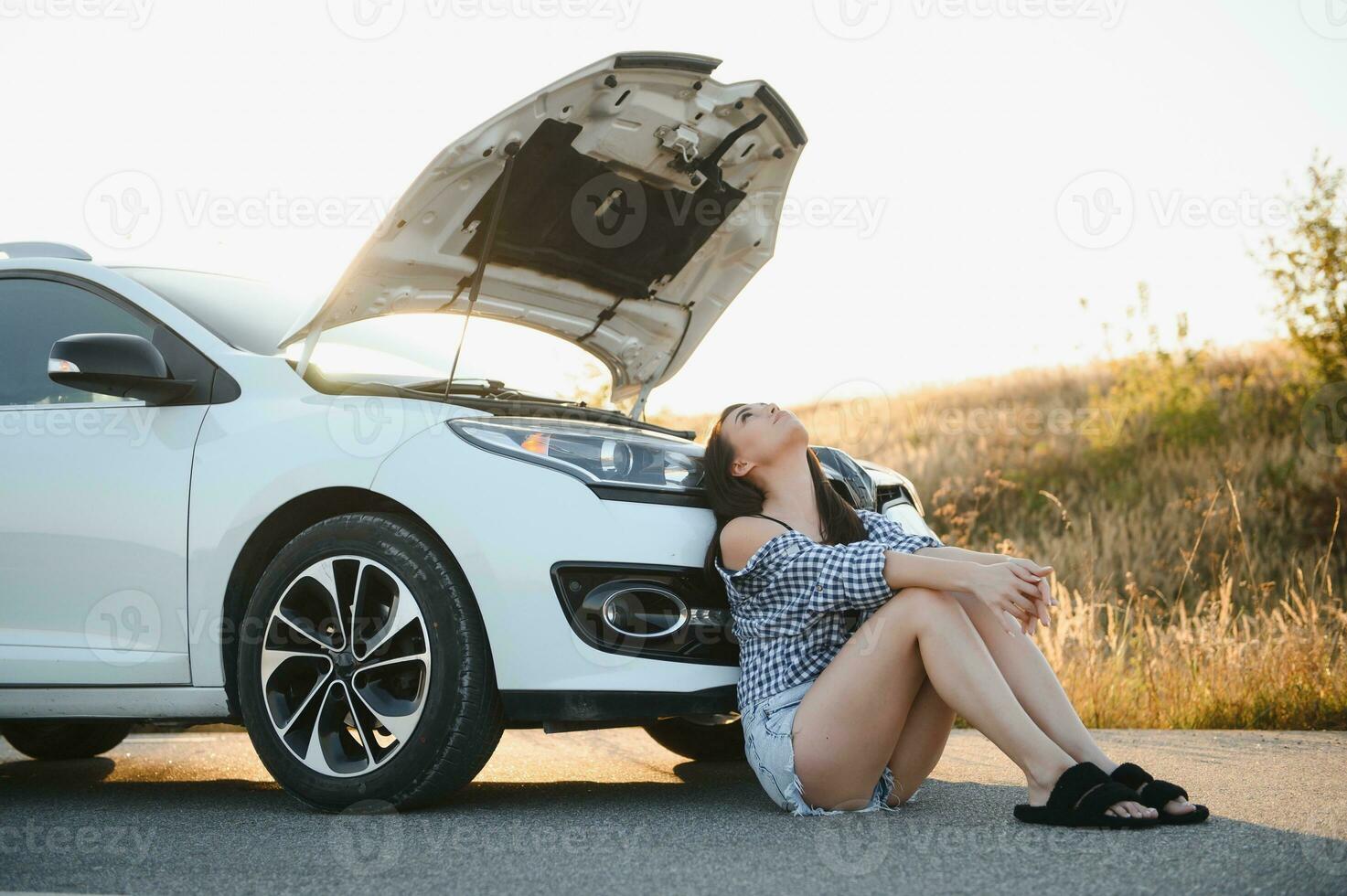 un' giovane ragazza si siede vicino un' rotto auto su il strada con un Aperto cappuccio. foto