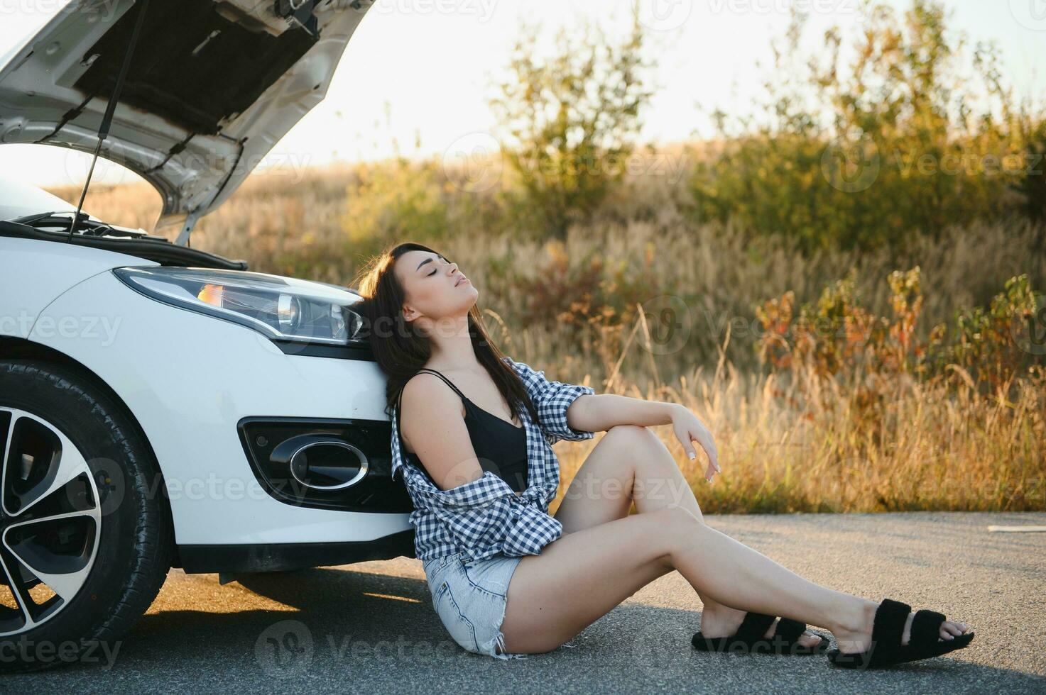 un' giovane ragazza si siede vicino un' rotto auto su il strada con un Aperto cappuccio. foto