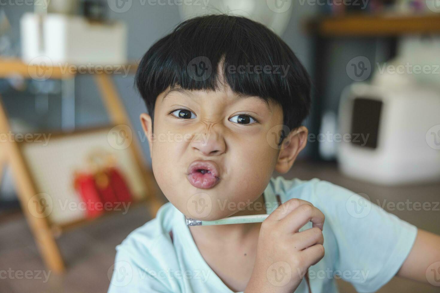 vicino su ritratto di asiatico ragazzo con nero scoppi, nero occhi con un' sorridente viso indossare un' leggero verde camicia Guarda a telecamera e seduta su il pavimento di il suo Casa e mano Tenere matita per disegno. foto