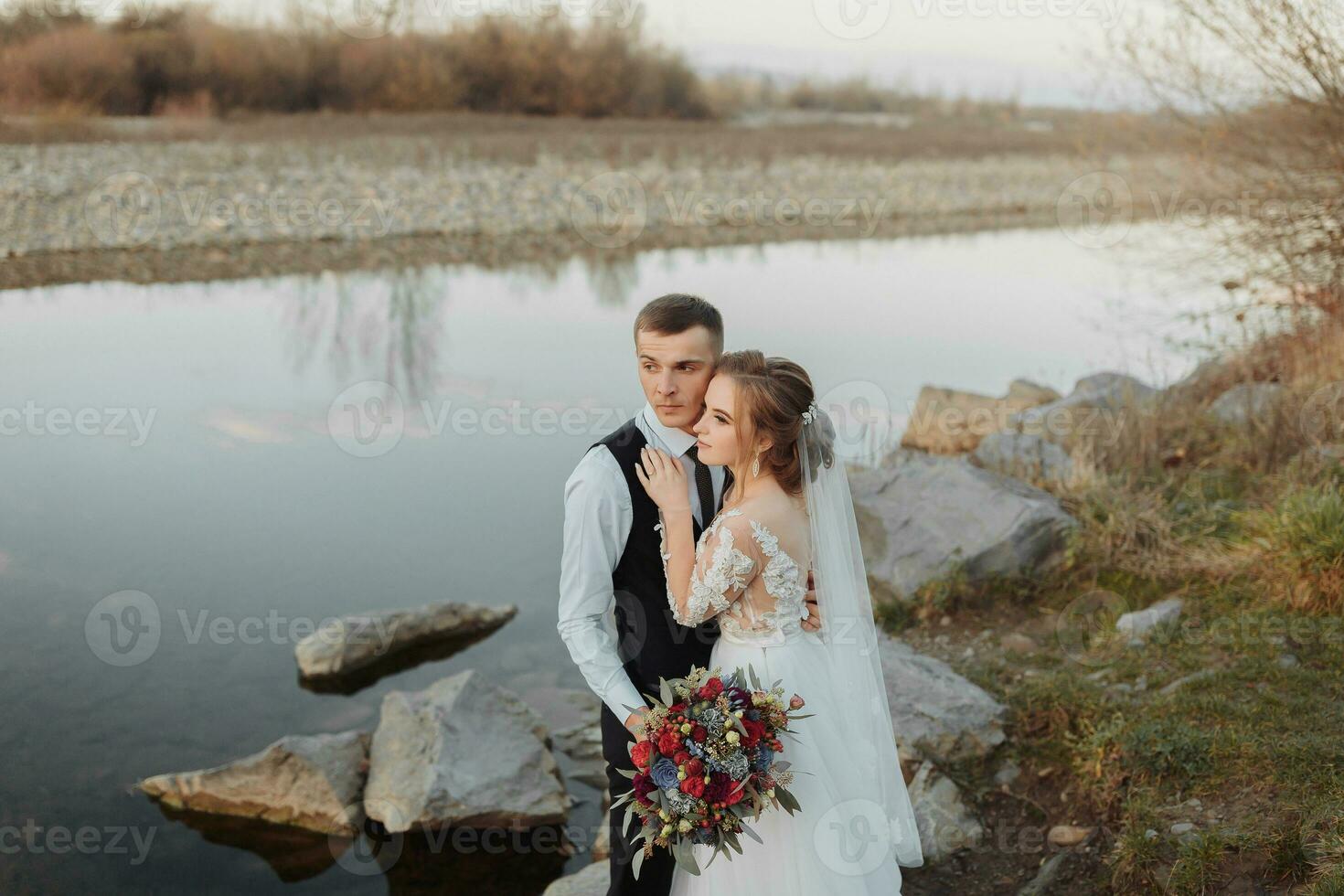 spose su il banca di il fiume, con spoglio gambe abbracciare e baci su un' roccia Tenere un' mazzo di vario fiori, rosso Rose e blu fiori e un' lungo bianca vestito foto