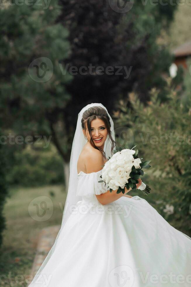 nozze foto. il sposa nel un' voluminoso bianca vestito e un' lungo velo, sorridente, volteggiare con un' mazzo di bianca Rose, Tenere sua vestire. ritratto di il sposa. bellissimo trucco e capelli. foto