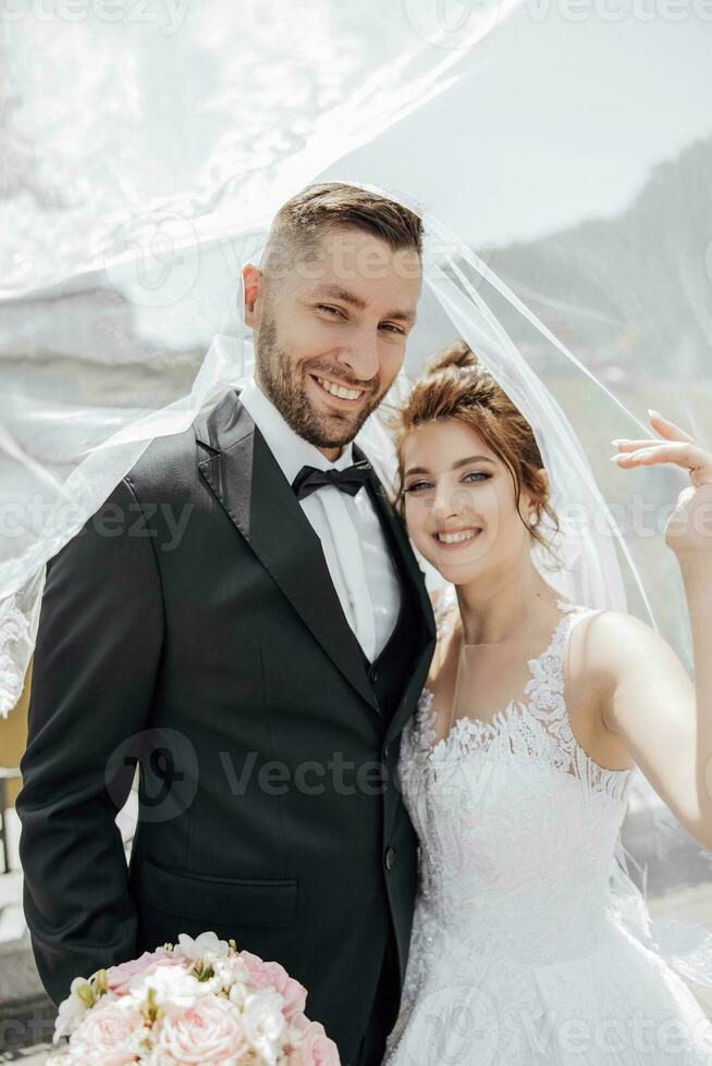 elegante bellissimo coppia sotto velo. abbracci su ogni Altro. essi Guarda in il telecamera e Sorridi Cordiali saluti. amore nel il occhi. bellissima leggero Aggiunge un' romantico atmosfera foto