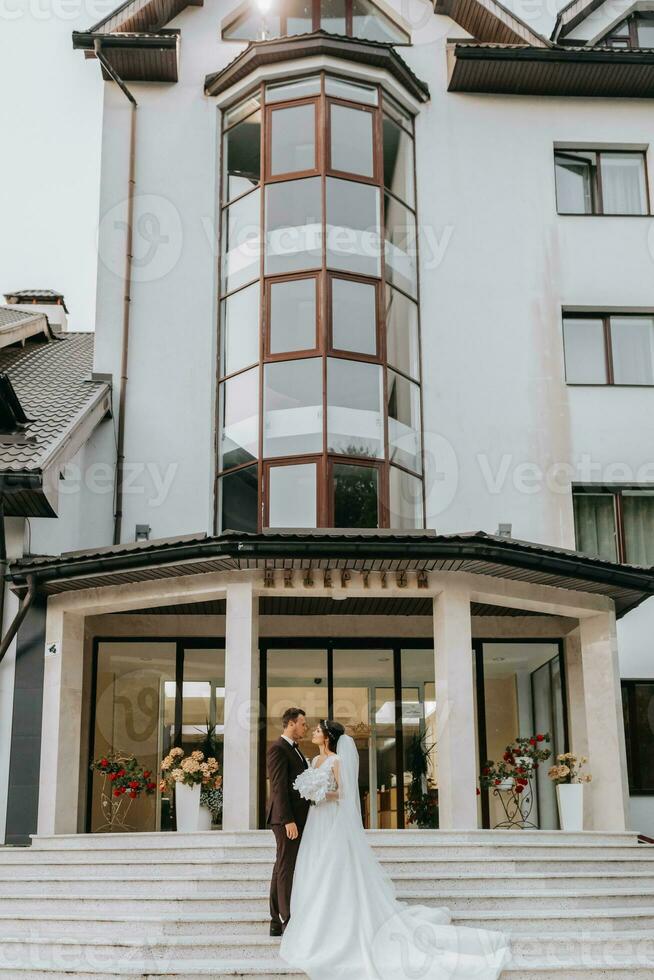 sposo e sposa a piedi all'aperto vicino un' elegante Hotel. un' lungo treno di vestito su il passaggi foto