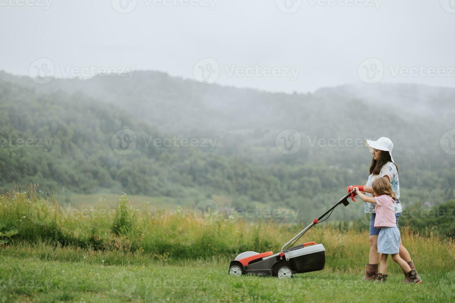 un' donna nel stivali con sua bambino nel il modulo di un' gioco falcia il erba con un' taglia erba nel il giardino contro il sfondo di montagne e nebbia, giardino utensili concetto foto