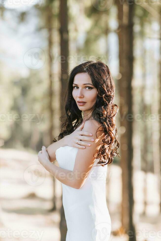 un' ragazza con lungo nero Riccio capelli soffiaggio nel il vento è in piedi nel un' bianca seta vestire. in piedi nel il primavera foresta contro il sfondo di grande alberi foto