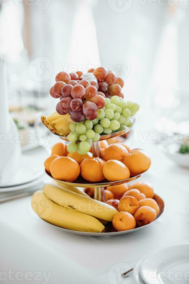 un' a tre livelli frutta piatto pieno con agrume frutta e uva sta su un' bianca tovaglia. primavera nozze foto