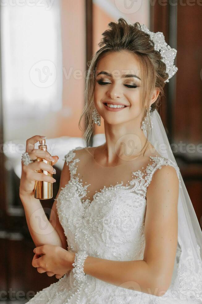 femmina bellezza. bello donna a casa. sposa spray profumo elegante donna indossare un' bianca vestito spray tenero profumo. elegante bicchiere bottiglia di profumo nel mani. ragazza con trucco e un' bottiglia di profumo foto